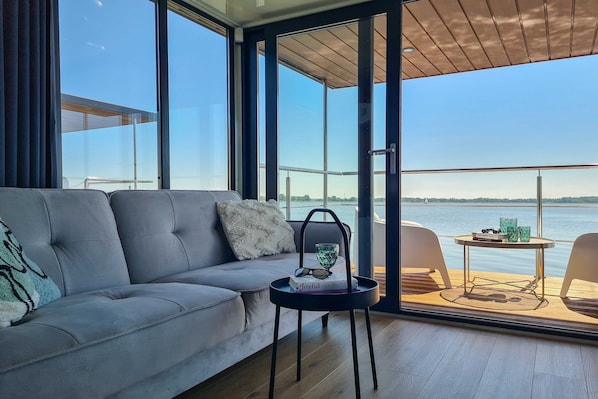 A modern living room with a sofa, a small table with a book and glasses, and a view of a body of water through large windows.