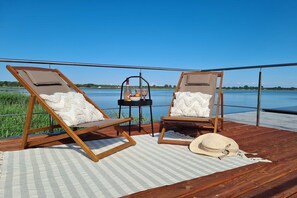 Dos sillones de madera con cojines, una mesita con una botella y fruta, y un sombrero de paja sobre una estera a cuadros en una terraza de madera con vista a un cuerpo de agua.