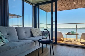 Un salon moderne avec un canapé, une petite table sur laquelle se trouvent un livre et des lunettes, et une vue sur une étendue d’eau grâce à de grandes fenêtres.