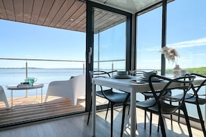 A dining area with a table and chairs, large windows with a view of the water, and a wooden deck outside. - Houseboat in Łazy by Lake Jamno and Sea (Łazy)