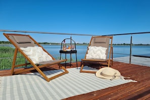 Two wooden lounge chairs with cushions, a small table with a bottle and fruit, and a straw hat on a checkered mat on a wooden deck overlooking a body of water. - Houseboat in Łazy by Lake Jamno and Sea (Łazy)