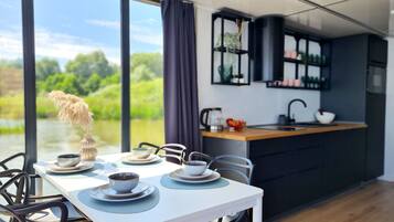 Un comedor moderno con una mesa preparada para dos, una cocina con armarios oscuros y vistas a la vegetación a través de amplias ventanas.