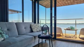 Ein modernes Wohnzimmer mit einer Couch, einem kleinen Tisch mit Buch und Gläsern und Blick auf ein Gewässer durch große Fenster.