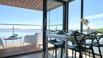 Un comedor con mesa y sillas, ventanas amplias con vista al agua y una terraza de madera afuera.