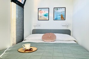 A neatly made bed with a green quilt, a pink pillow, and a tray with a mug and snacks. There are two framed pictures on the wall, one depicting a beach scene and the other a person jumping off a cliff.