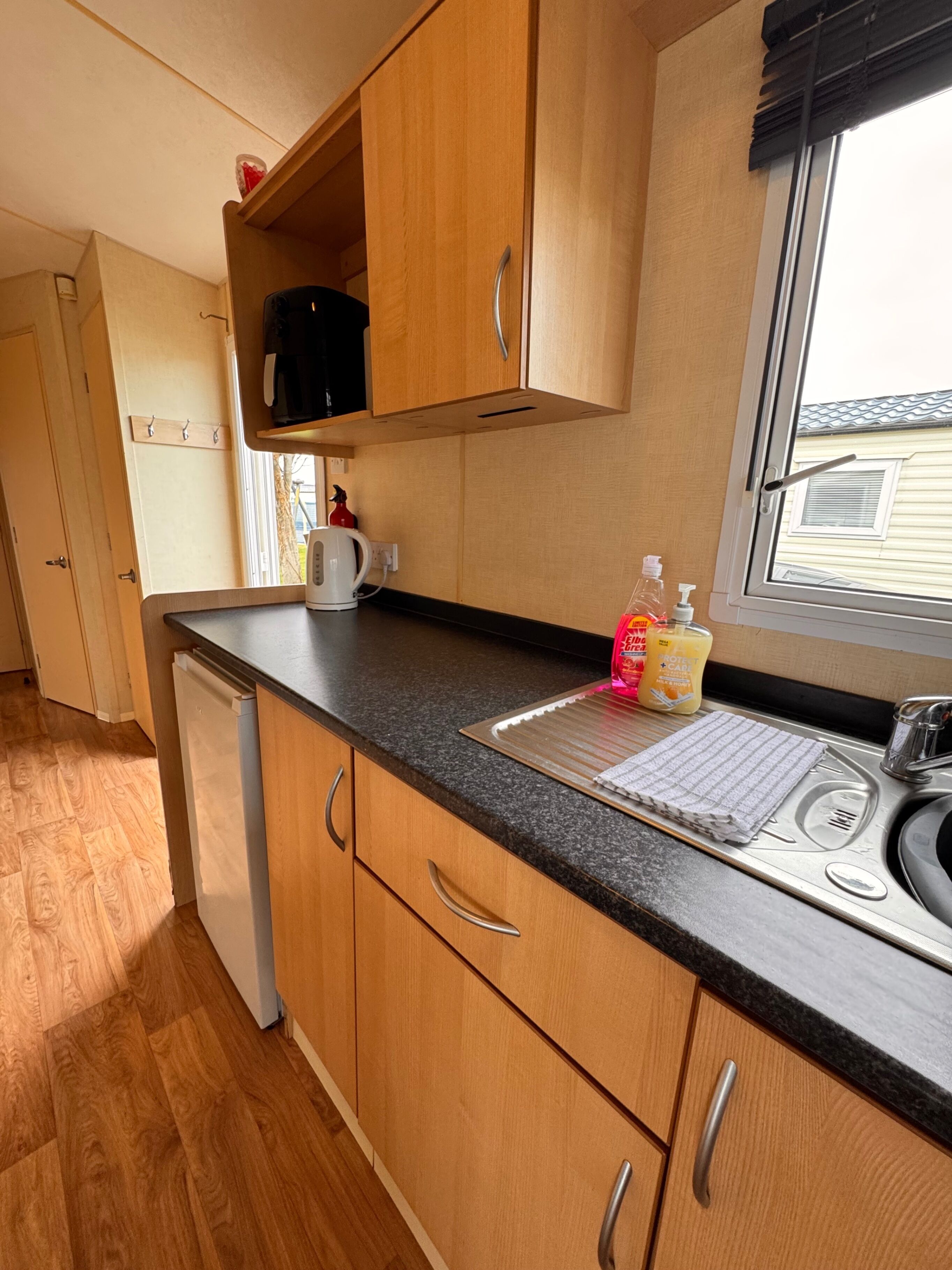 Ett kompakt kök med träskåp, en inbyggd mikrovågsugn, en vattenkokare och ett fönster med utsikt över ett hus.