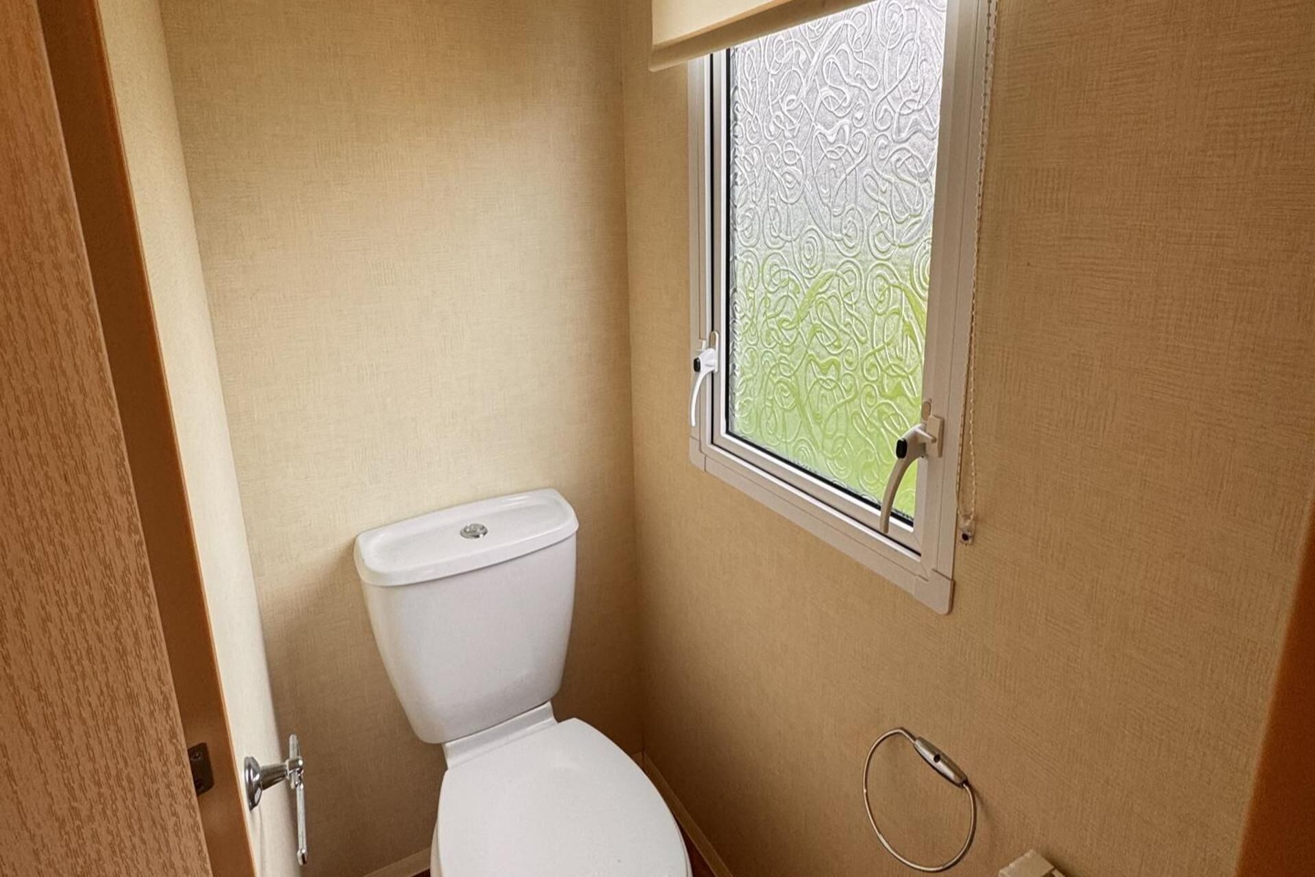 Casa de banho pequena e compacta, com sanita, janela com vidro jateado e suporte para toalha na parede.