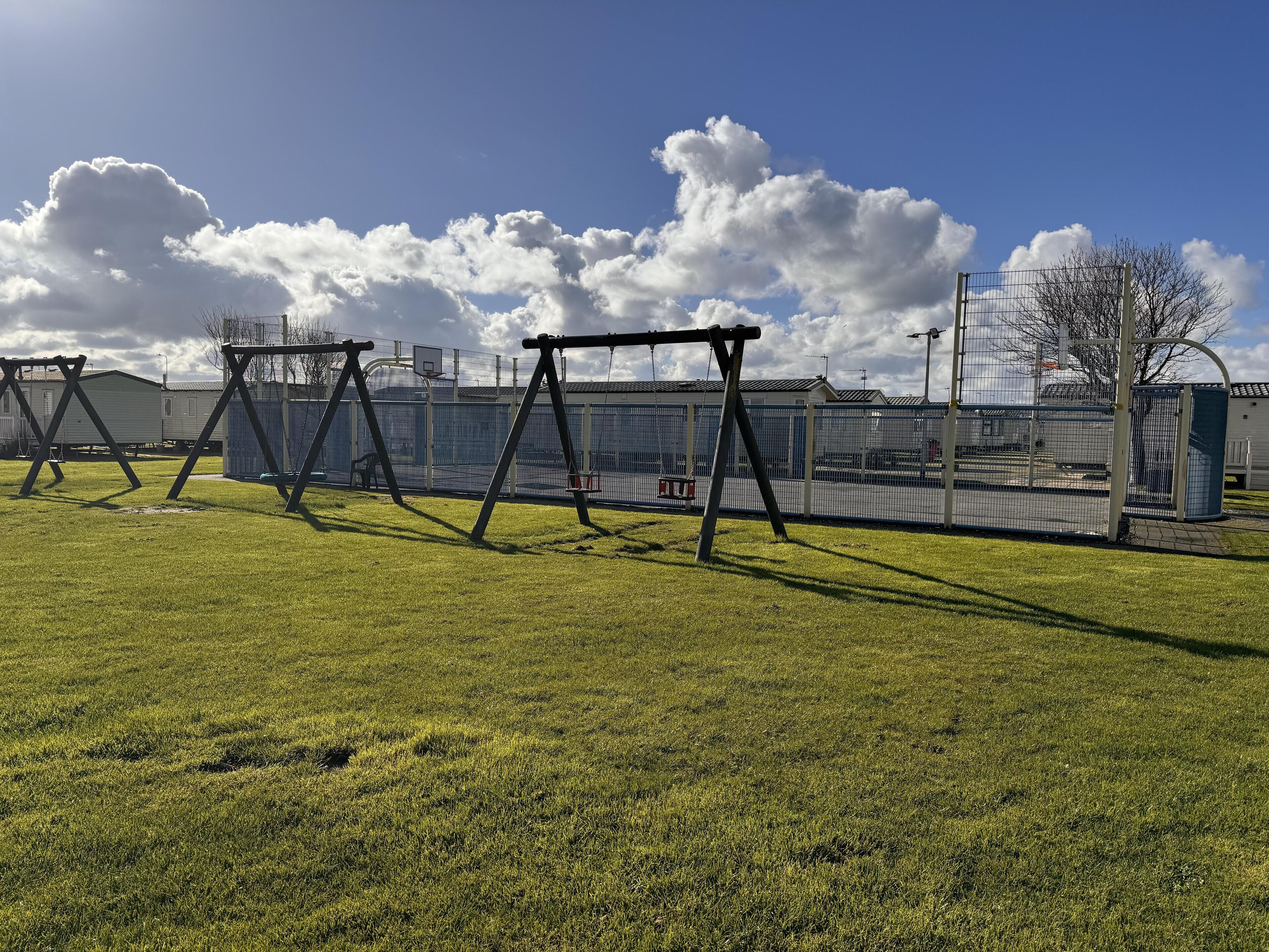 A grassy outdoor area with swings and a fenced basketball court.