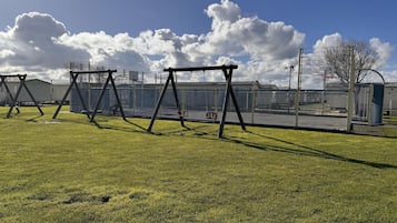 A grassy outdoor area with swings and a fenced basketball court.