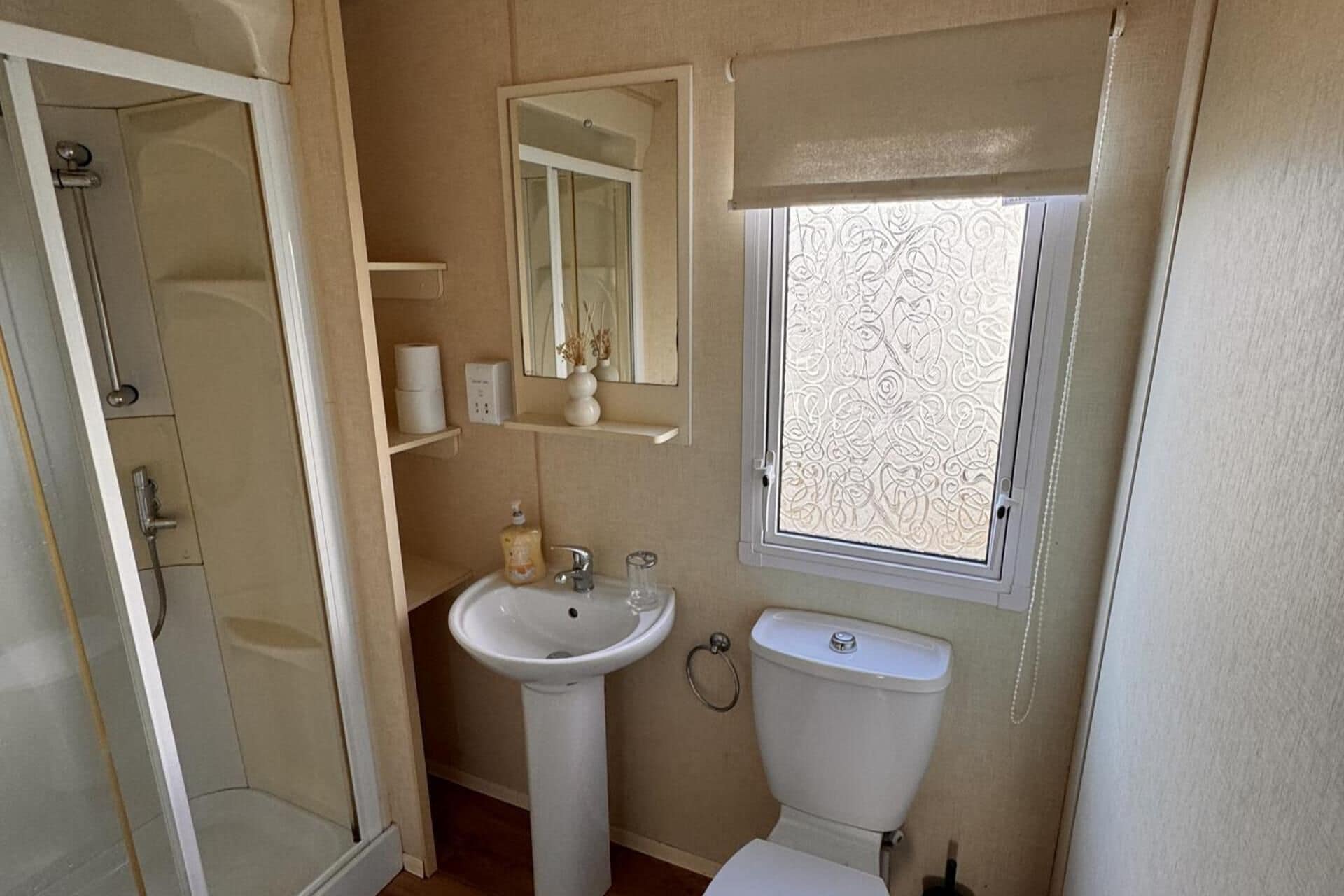 A compact bathroom with a shower, sink, toilet, and a window with decorative blinds.
