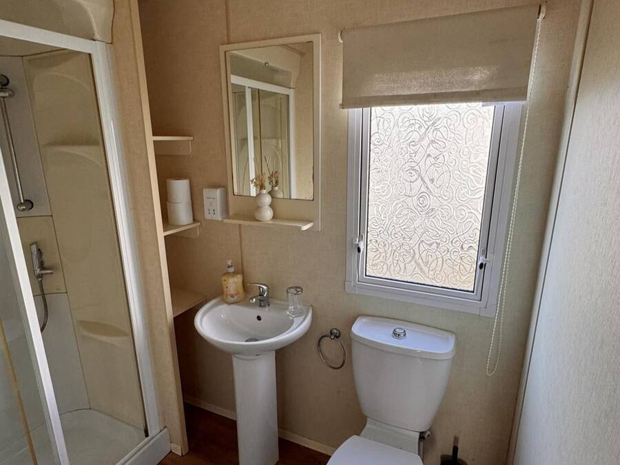 A compact bathroom with a shower, sink, toilet, and a window with decorative blinds.