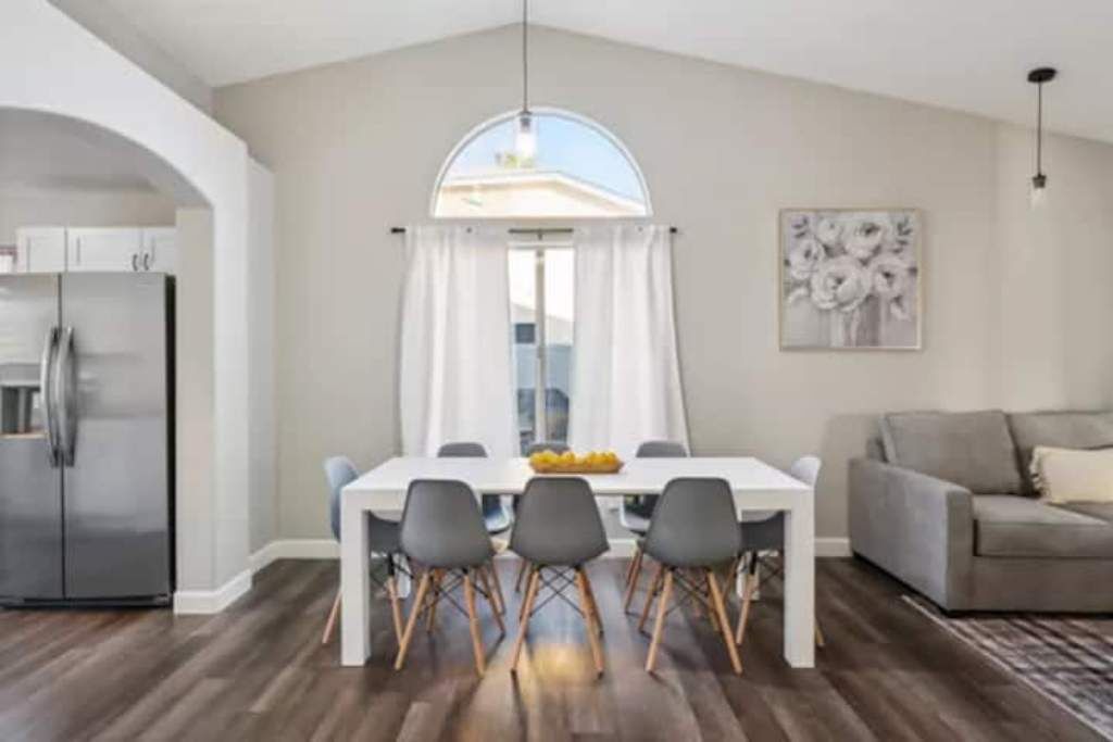 A modern dining area with a white table and chairs, a large window with curtains, a grey sofa, and a kitchen with a refrigerator.