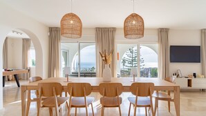 Ein moderner Essbereich mit einem langen Tisch und sechs Holzstühlen, großen Fenstern mit Vorhängen und Blick ins Freie.