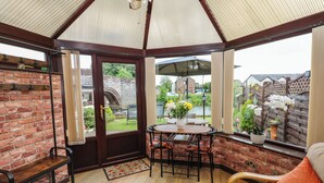 A cozy room with a brick wall, a wooden bench, a table with chairs, and a view of a garden through a glass door. - River Cottage (Penrith)