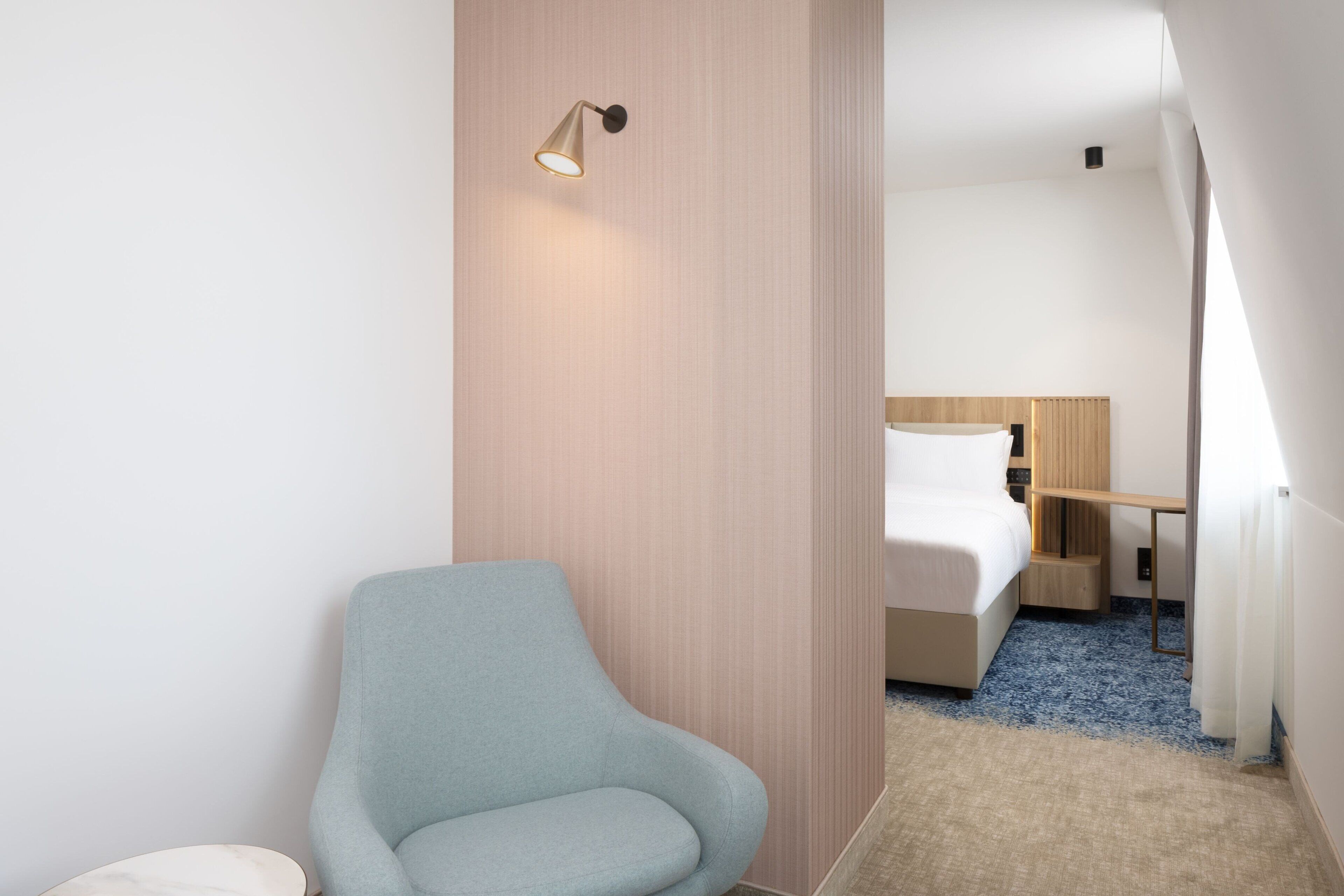 Habitación de hotel moderna con un sillón azul claro, una cama con cabecero de madera, un escritorio pequeño y un radiador.