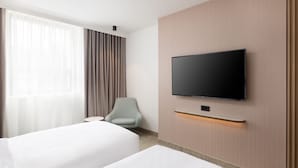 Ein Hotelzimmer mit zwei Betten, einem an der Wand montierten Fernseher, einem Sessel und einem Fenster mit Vorhängen.