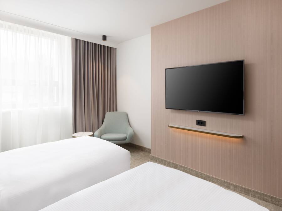 A hotel room with two beds, a television mounted on the wall, a chair, and a window with curtains.