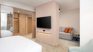 A modern hotel room with a large bed, a TV mounted on a wooden panel, a sofa with orange cushions, and a small table.