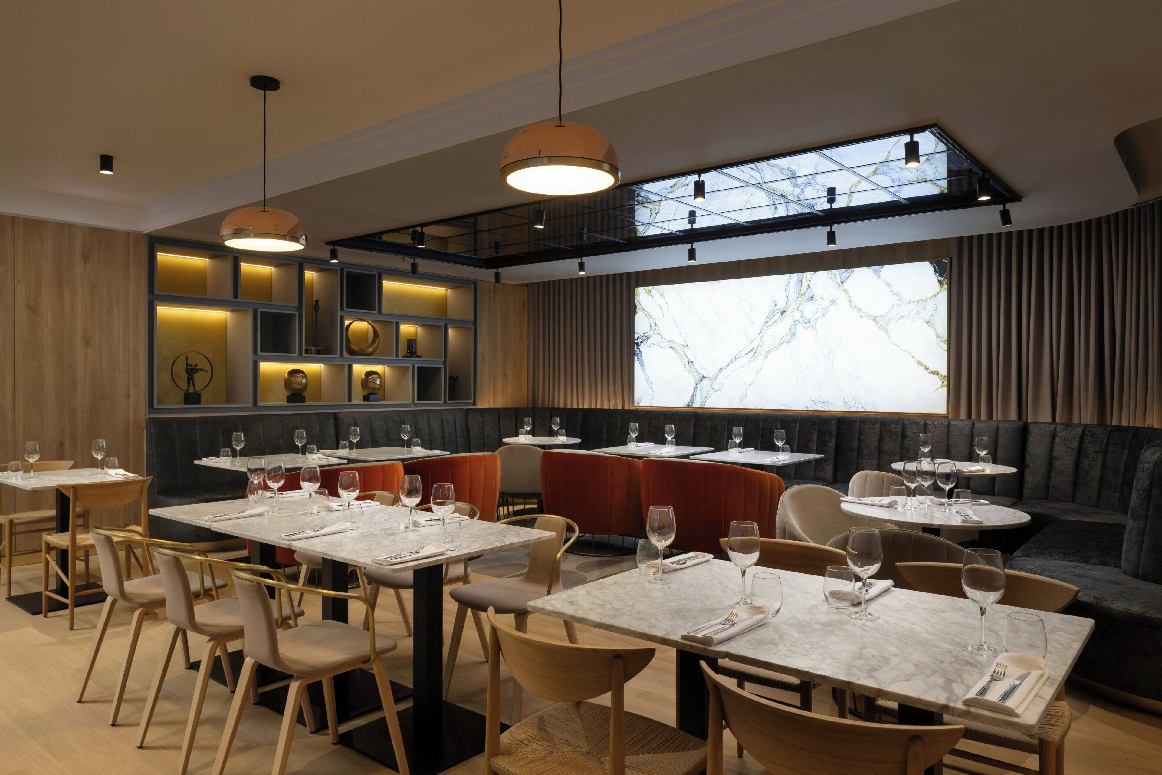 A modern restaurant interior with multiple dining tables, cushioned seating, and a large window providing natural light.