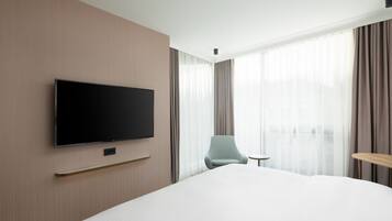 A modern hotel room with a large bed, a flat-screen TV mounted on the wall, a small round table, a chair, and a window with sheer curtains.