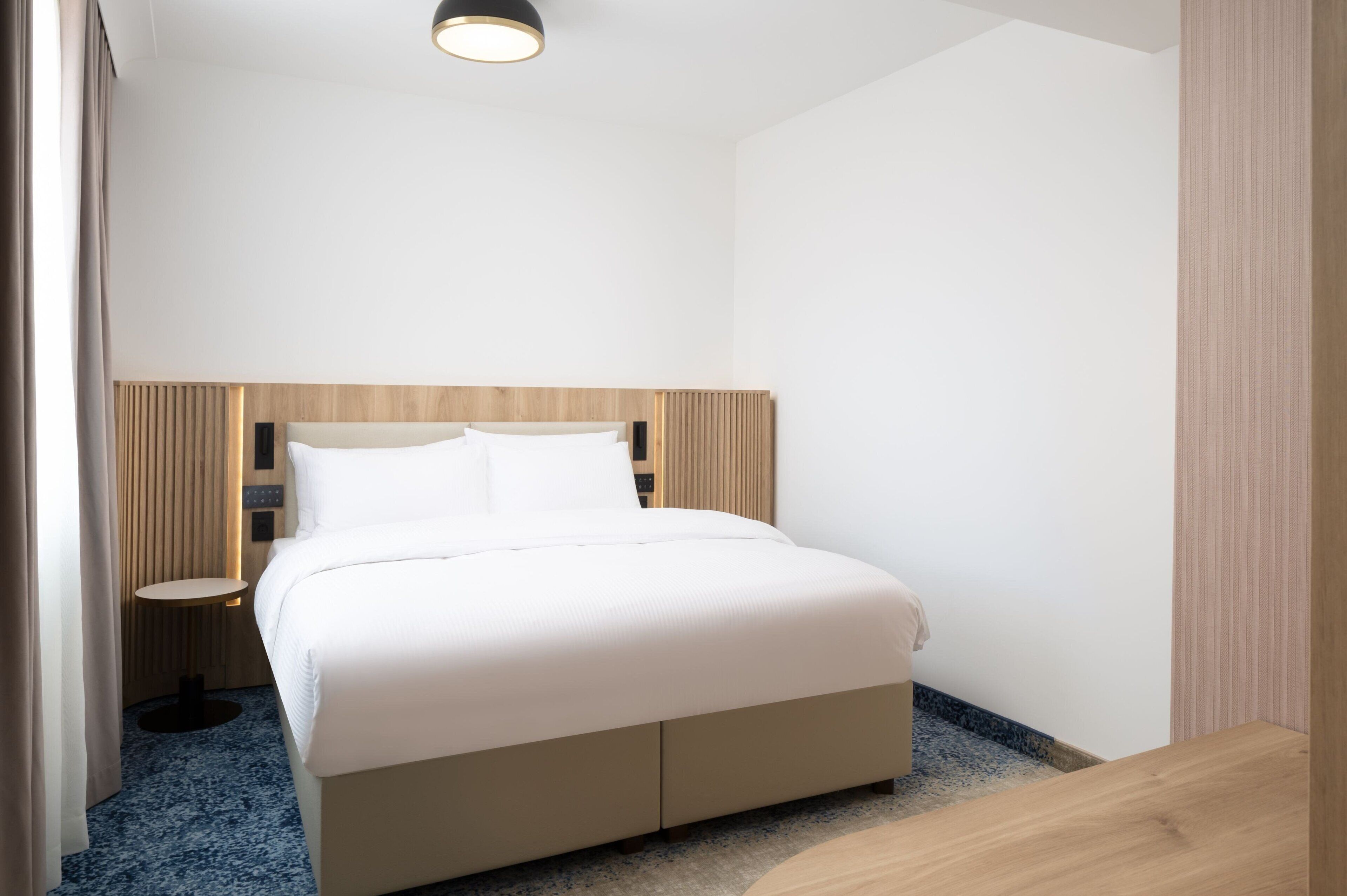 A modern hotel room with a large bed, a bedside table, and a small round table.