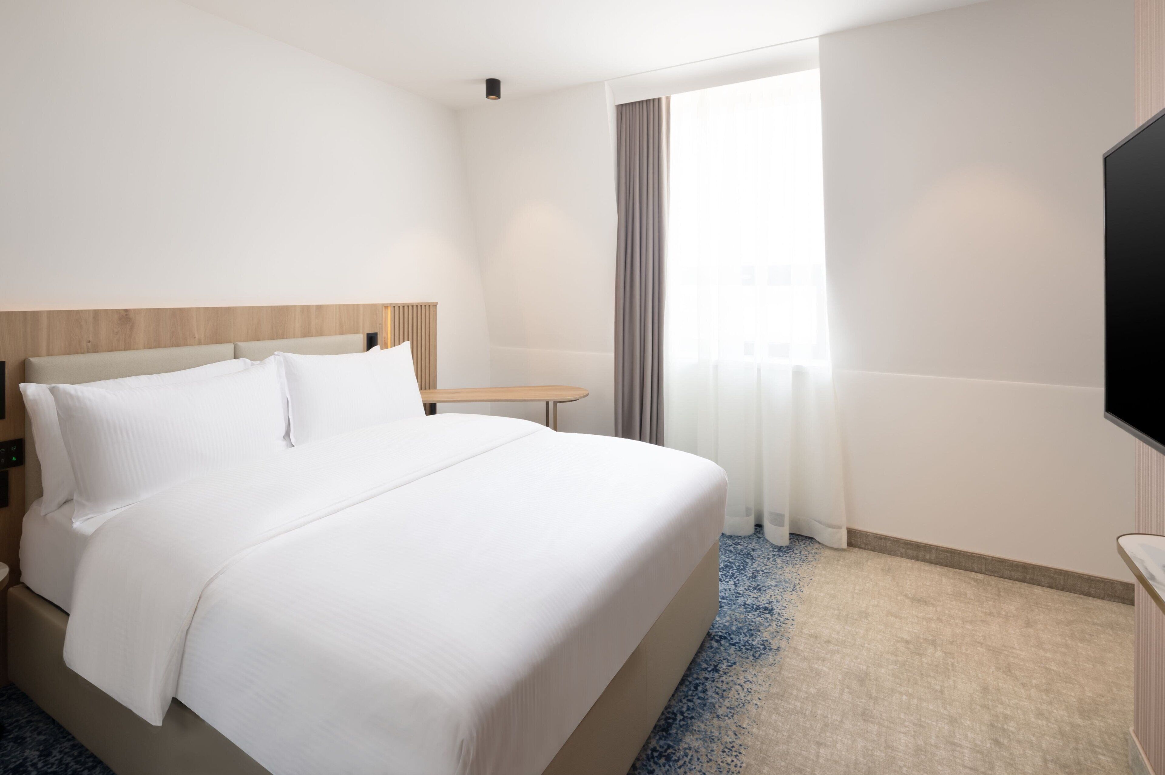 Quarto de hotel com uma cama grande, mesa de cabeceira, televisão de tela plana e janela com cortinas.