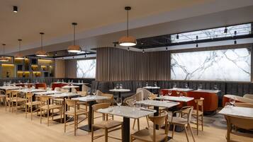 A modern restaurant with multiple dining tables and chairs, large windows with a view, and pendant lighting.