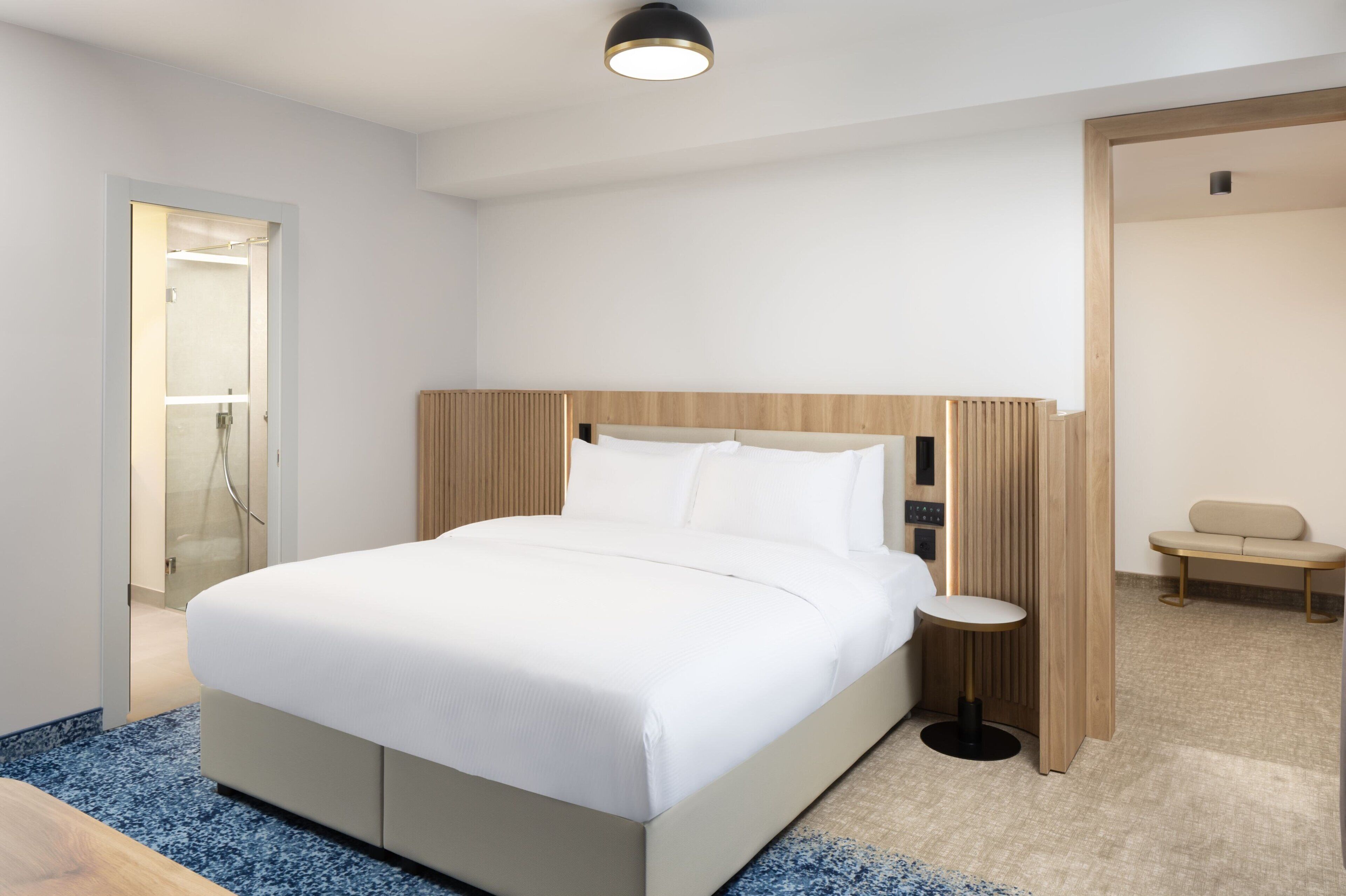 A modern hotel room with a large bed, a bedside table, a small round table, and a bench in the corner.