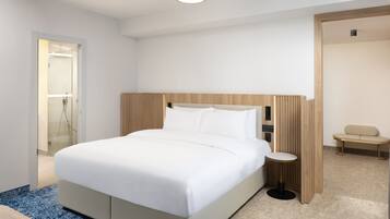 A modern hotel room with a large bed, a bedside table, a small round table, and a bench in the corner.