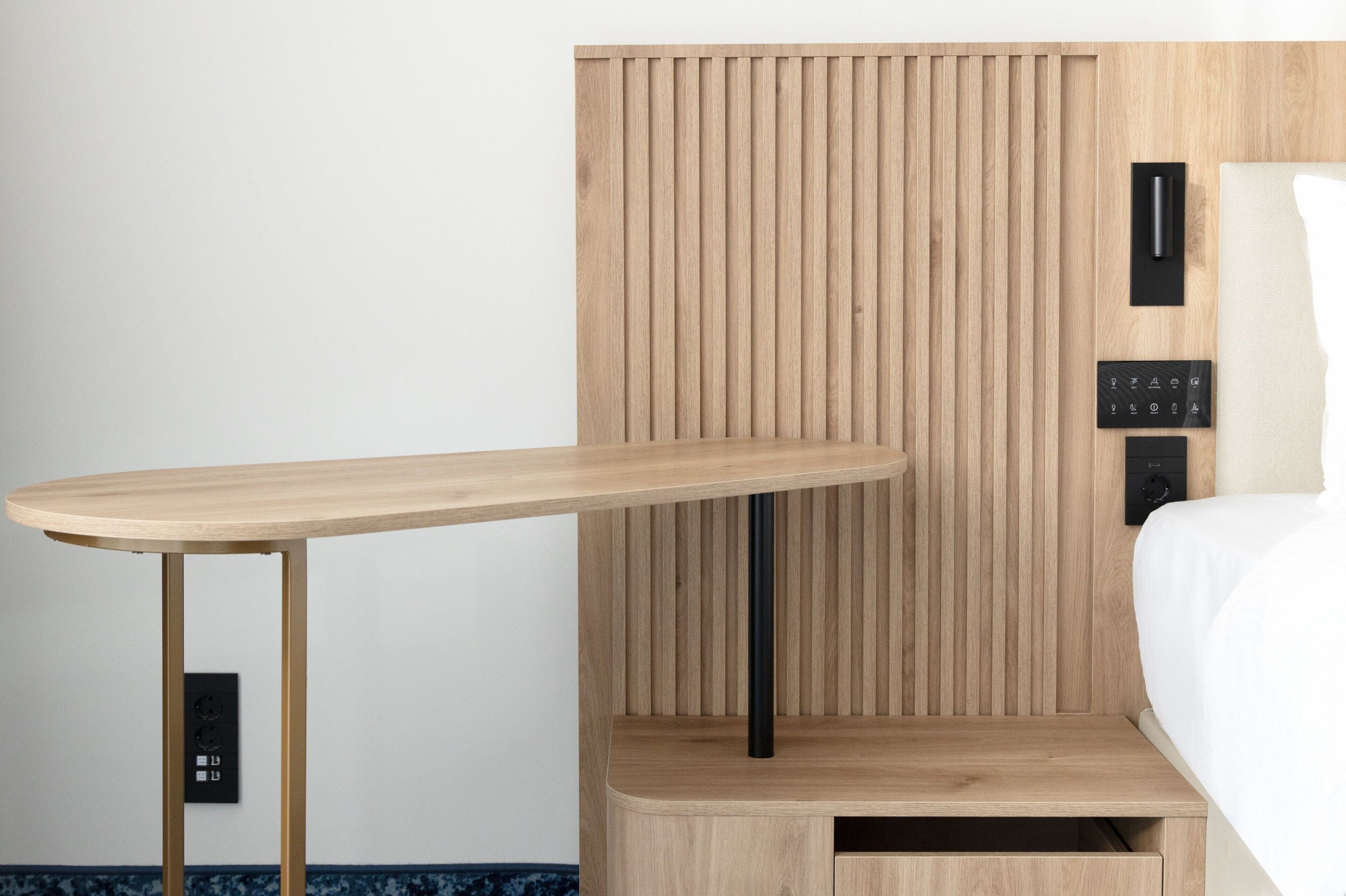 A modern room with a wooden bedside table, a built-in bedside console with a control panel, and a bed with white bedding.