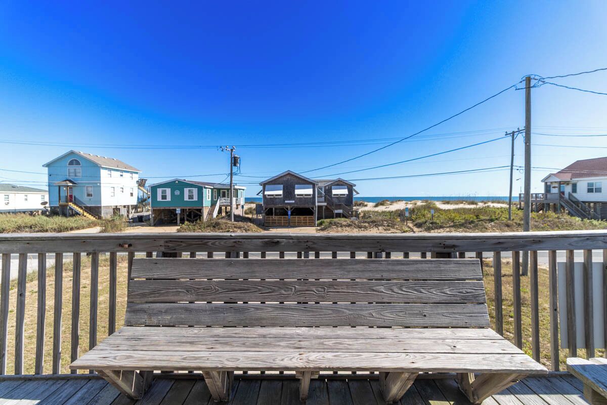 A wooden deck with a picnic table, overlooking a beach with houses and a clear blue sky.
