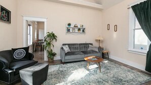 Ein Wohnzimmer mit grauem Sofa, einem schwarzen Ledersessel, einem Couchtisch und einer großen Pflanze. Im Hintergrund ist ein Essbereich zu sehen.