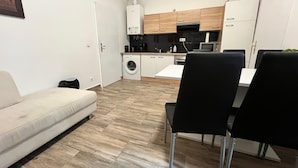 Un espace de vie compact comprenant un canapé, une table à manger avec des chaises, un lave-linge, un four à micro-ondes et un petit coin cuisine avec des placards en bois et un réfrigérateur blanc.