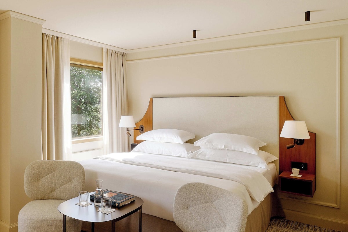 A hotel room with a large bed, two armchairs, a small table, and a nightstand with a lamp.