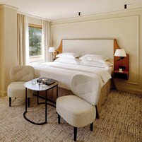 A hotel room with a large bed, two armchairs, a small table, and a nightstand with a lamp.