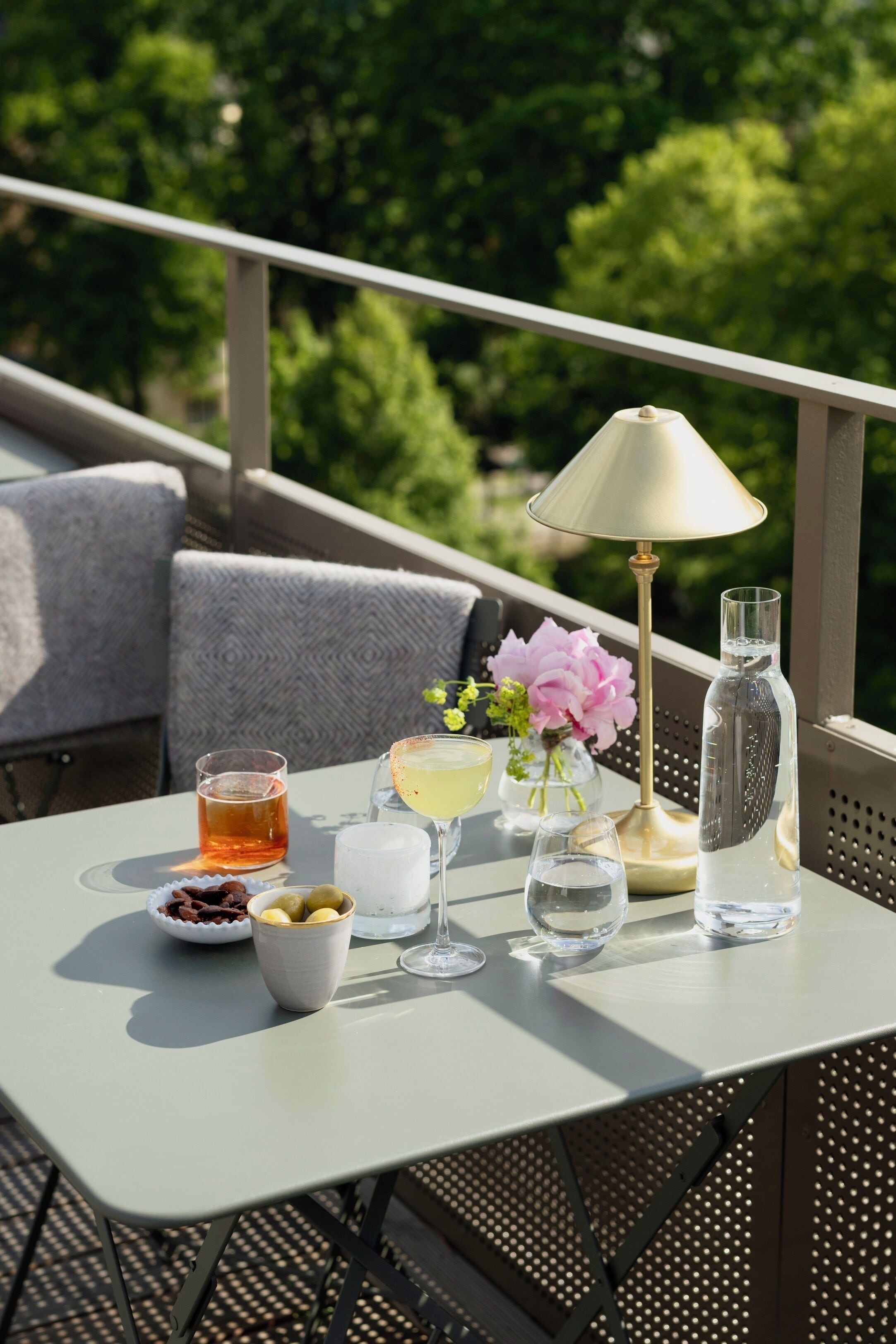 A table set outdoors with a table lamp, a bottle of water, a glass of iced tea, a bowl of chocolate, and a vase with flowers.
