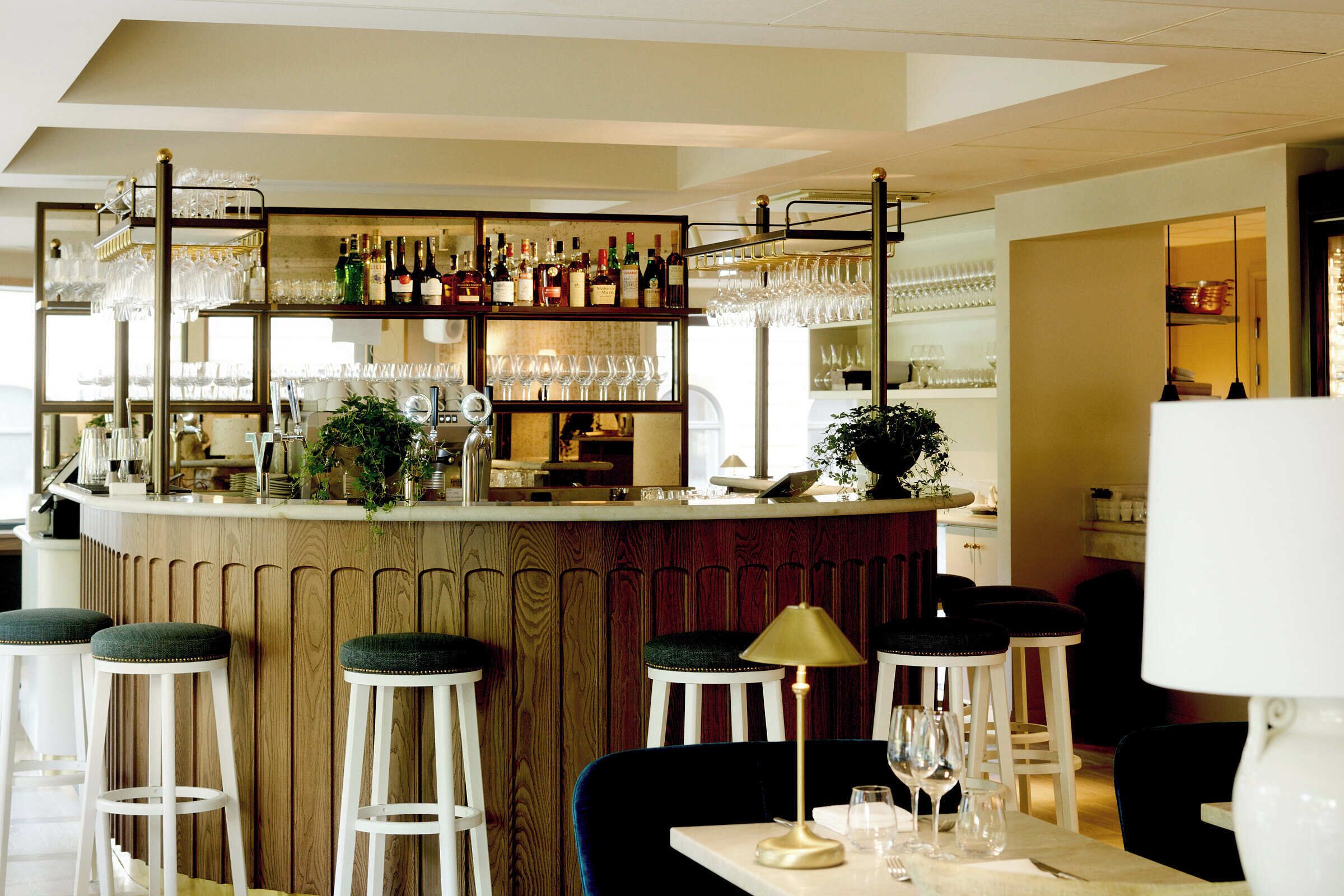 Zona de bar con taburetes, una barra con botellas y vasos, y una mesa con lámpara y copas de vino.
