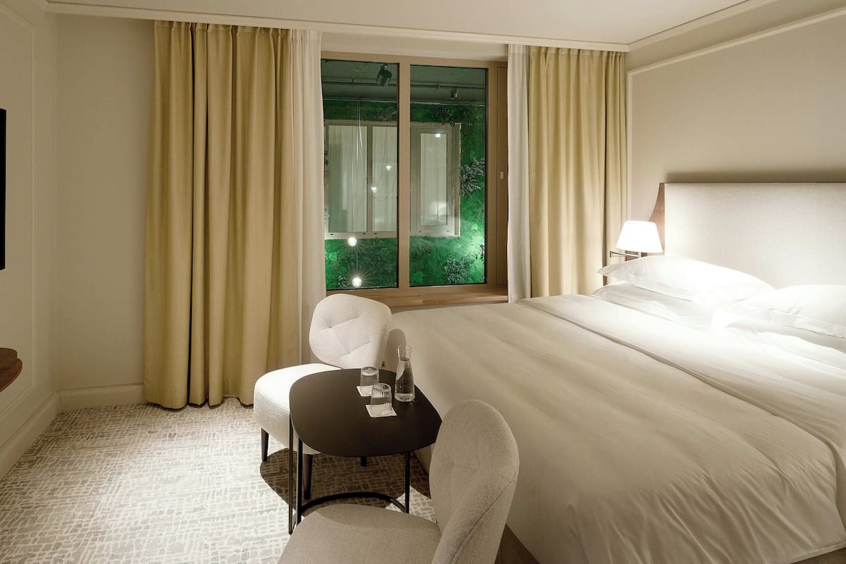 A hotel room with a large bed, a small table with a chair, a window with curtains, and a lamp on the bedside table.
