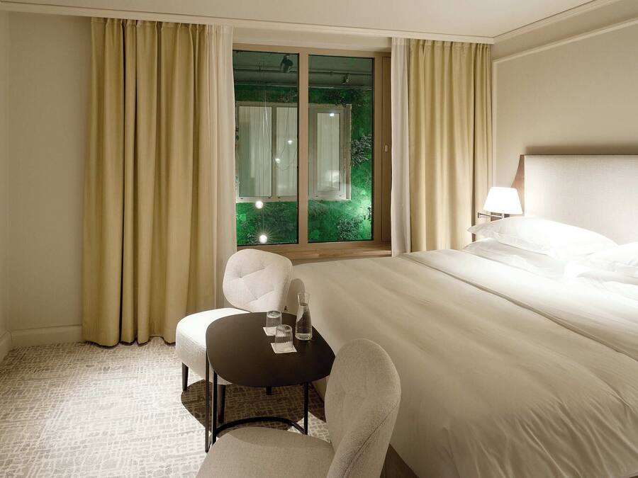 A hotel room with a large bed, a small table with a chair, a window with curtains, and a lamp on the bedside table.