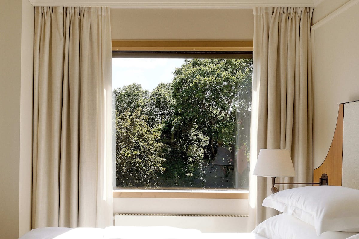 A hotel room with a large bed, a window with curtains, and a view of trees outside.