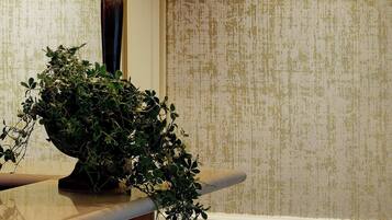Una lámpara con pantalla blanca sobre una superficie de madera, una planta en maceta y una pared con textura.