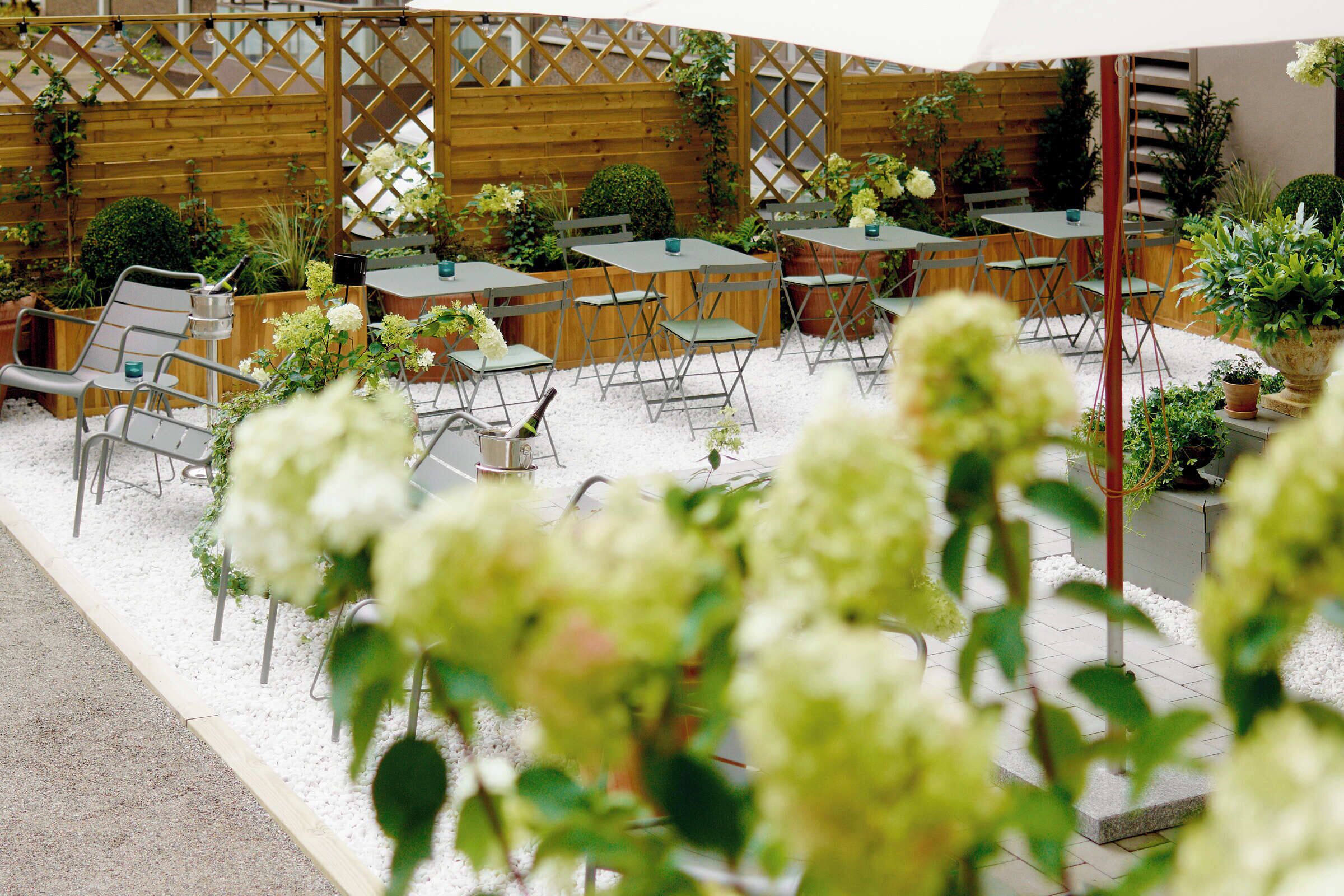 Un patio al aire libre con mesas y sillas, rodeado de plantas y una valla de madera.