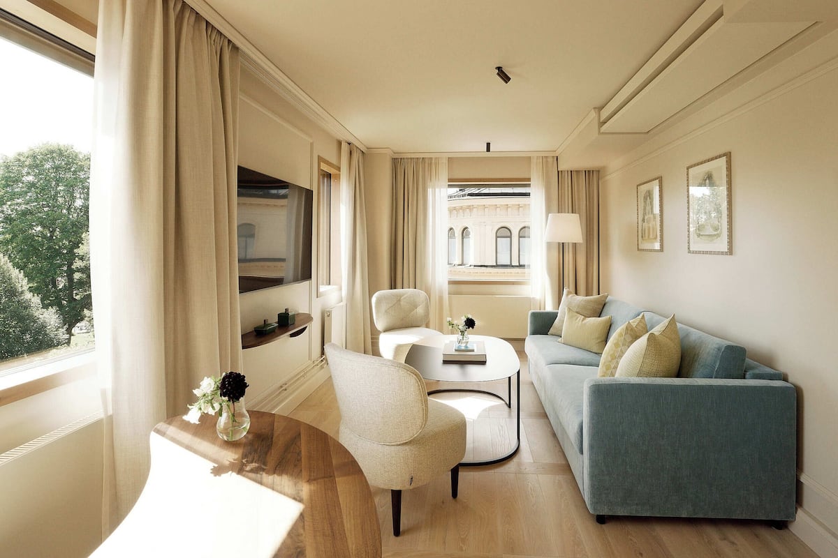 A modern living room with a blue sofa, a round coffee table, and a beige armchair. A window with curtains allows natural light to enter.