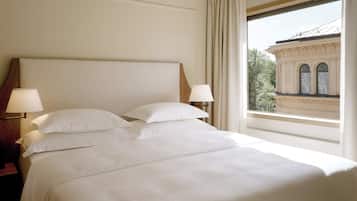 A neatly made bed with white linens and two bedside lamps, a window with a view of a building, and a wooden nightstand with a lamp.