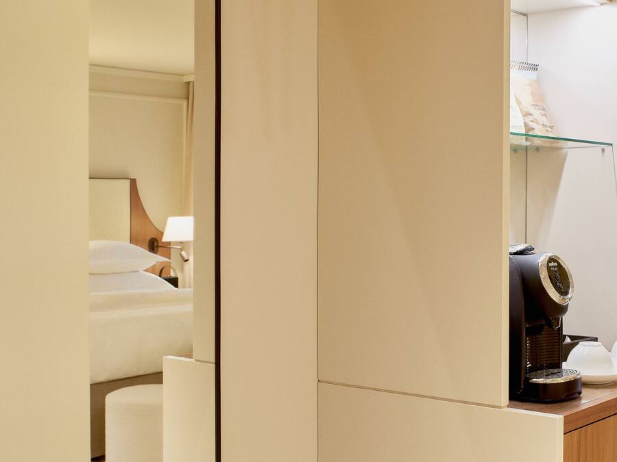 Ett hotellrum med ett torkställ av trä, en säng med vita sängkläder, en kaffemaskin och en hylla av glas med burkar.