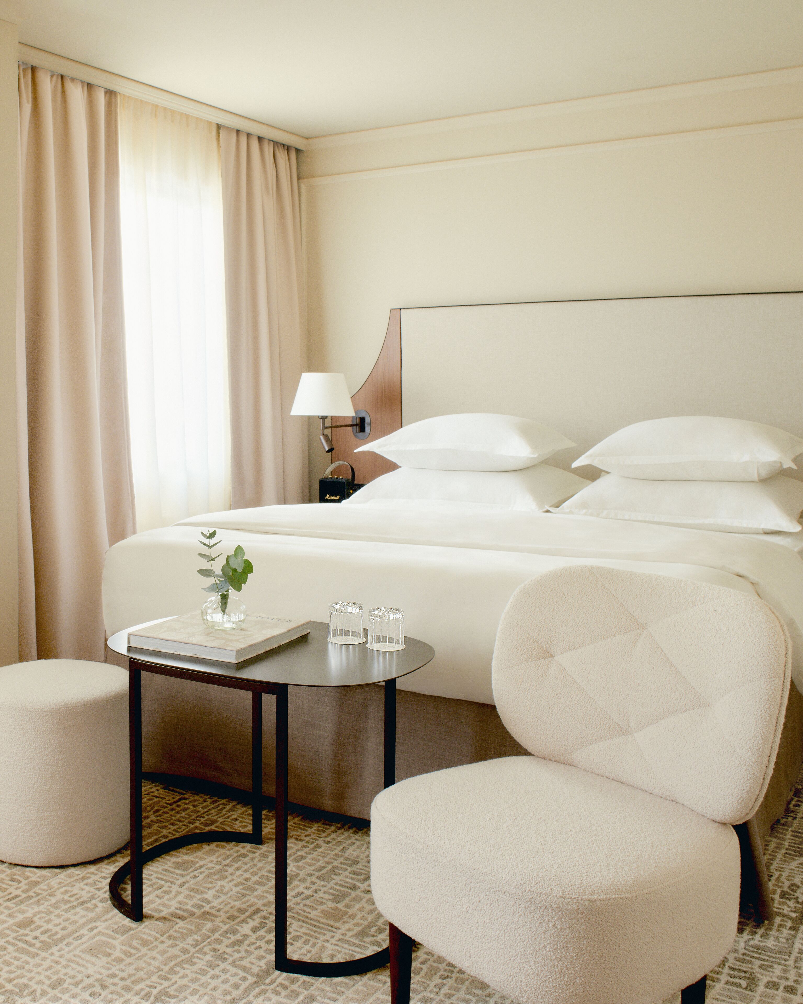 A hotel room with a bed, a bedside table with a lamp, a small table with a book and glasses, and a chair.