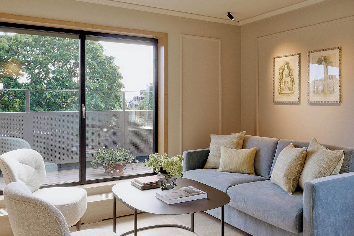 A modern living room with a grey sofa, white armchairs, and a round coffee table. There are two framed artworks on the wall and a balcony with a view of trees and buildings.