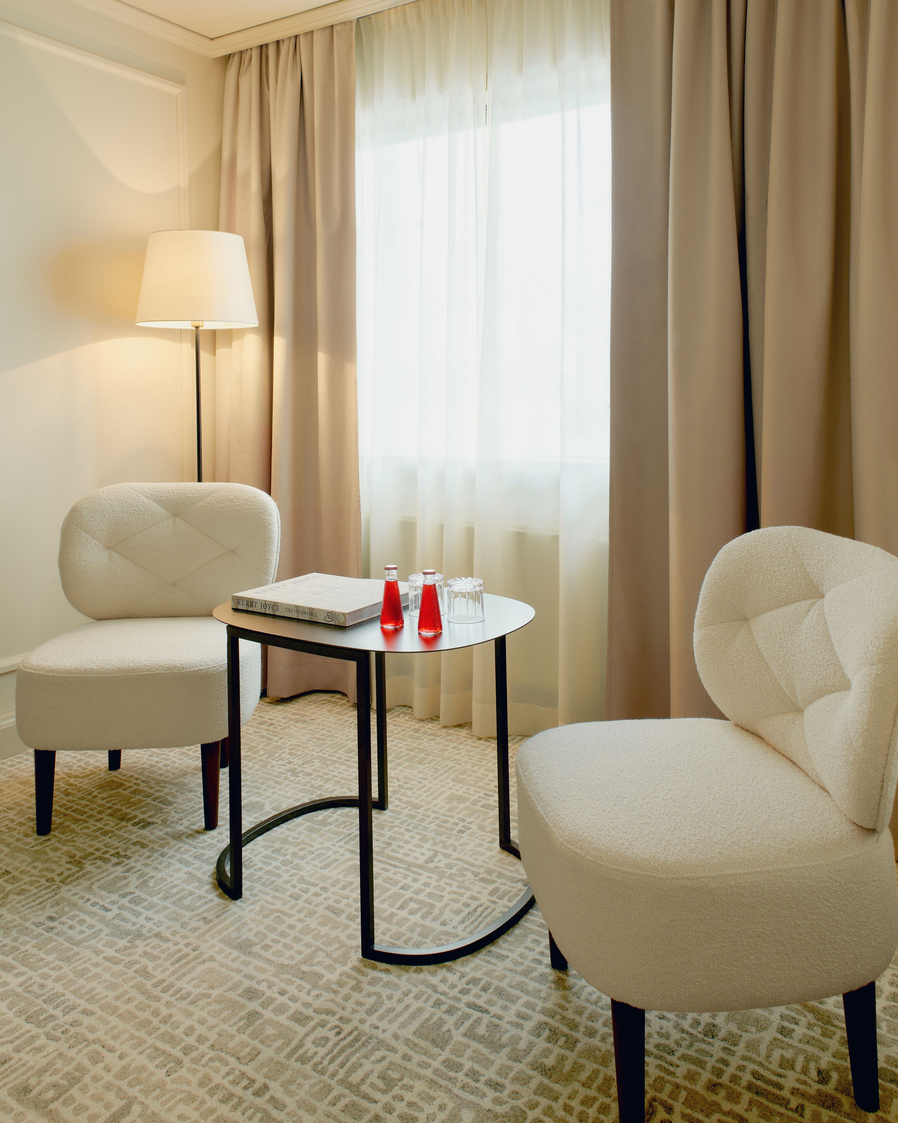 Une chambre d’hôtel avec deux fauteuils, une petite table sur laquelle se trouve un livre et deux bouteilles rouges, une lampe et une fenêtre ornée de rideaux légers.