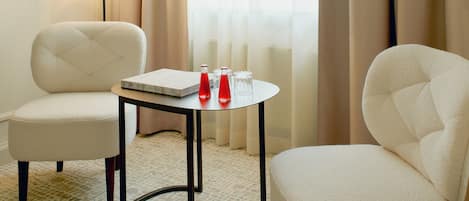 A hotel room with two armchairs, a small table with a book and two red bottles, a lamp, and a window with sheer curtains.