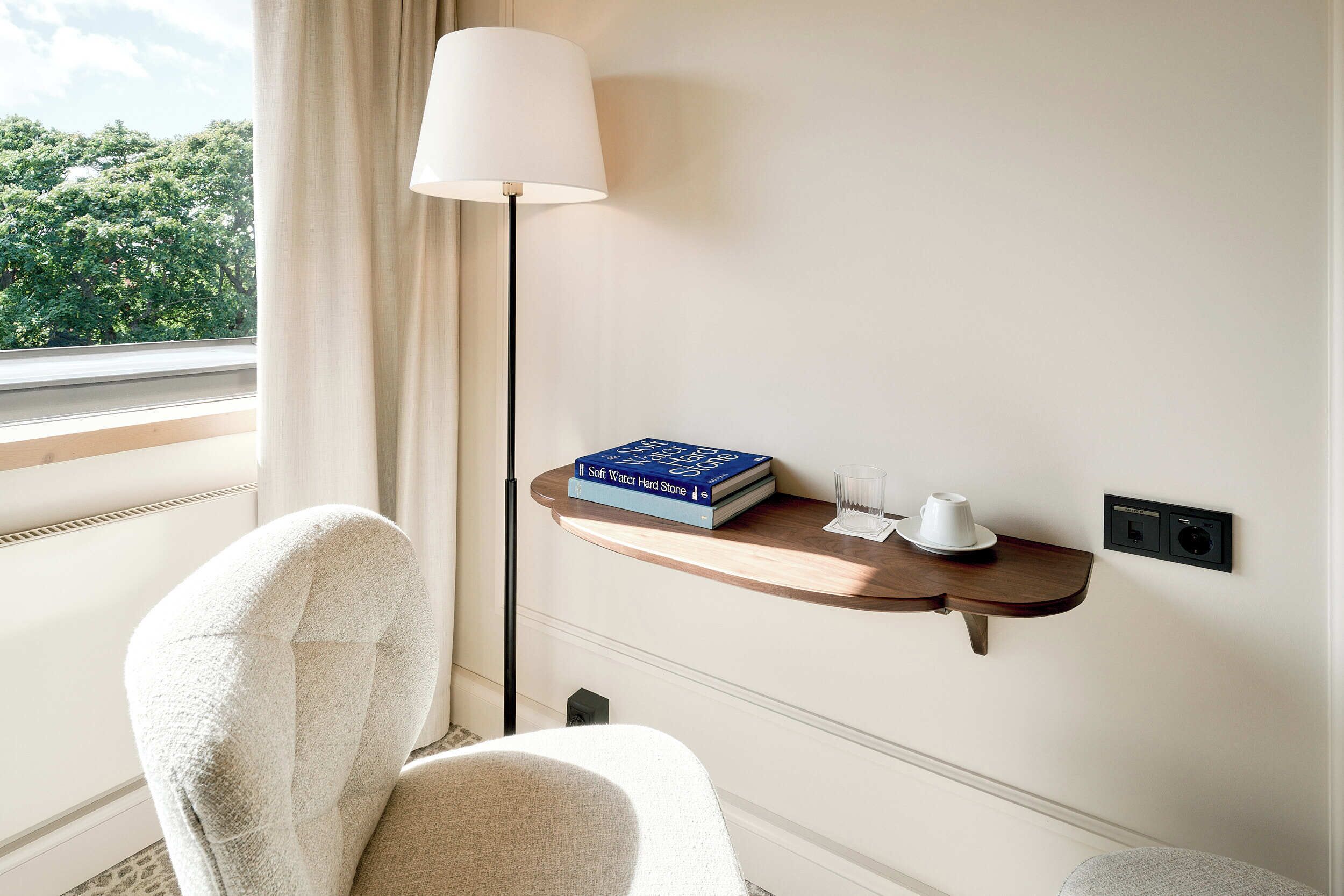 Une chambre d’hôtel avec une fenêtre, une chaise, une table de chevet avec un livre et un verre, et une lampe.